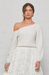 A woman is wearing a white, off-the-shoulder knit sweater with long sleeves. She has a matching white lace skirt that features intricate lace patterns. She accessorizes with large hoop earrings and rings on her fingers.